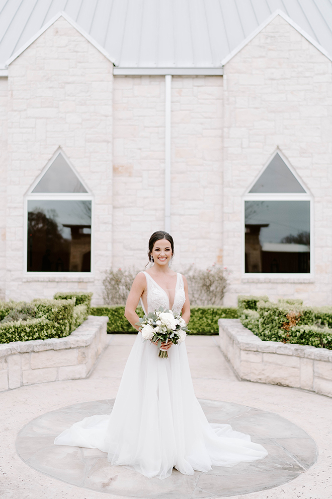 A bride in a v-neck wedding gown holds a small bouquet of white florals outside of a chapel at Briscoe Manor. 