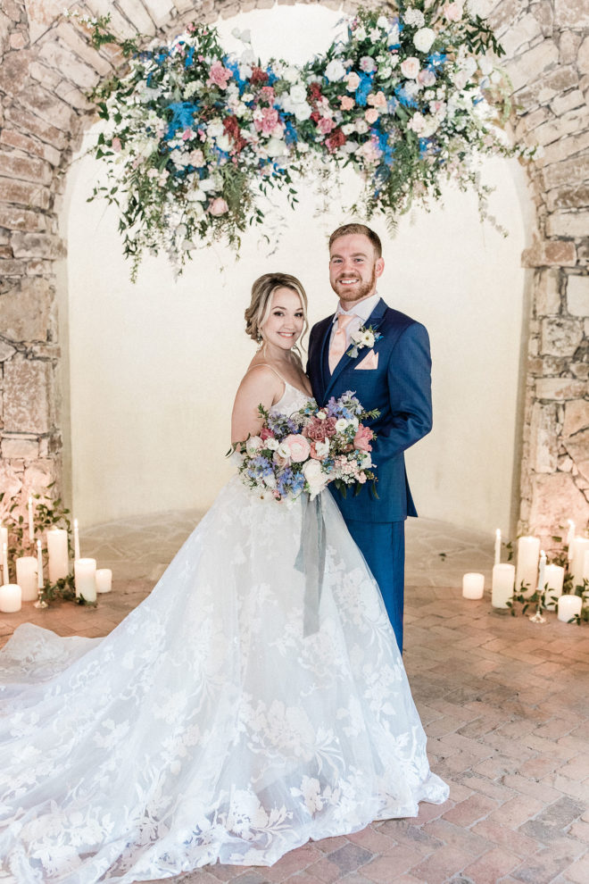 Bride and groom standing together a midst a pastel colored hanging floral installation set against brick arch inside of Camp Lucys Ian Chapel and glowing white candles surrounded by greenery.