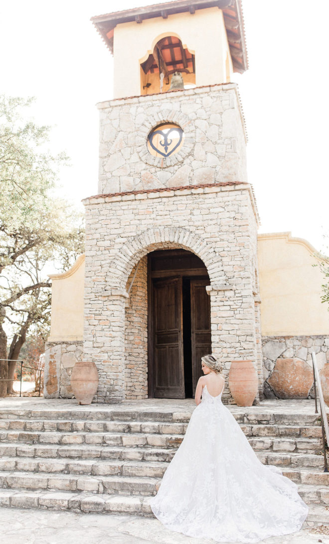 Bride standing on steps of light bright old world architecture of Ians Chapel at Camp Lucy with her back to camera and long lace train