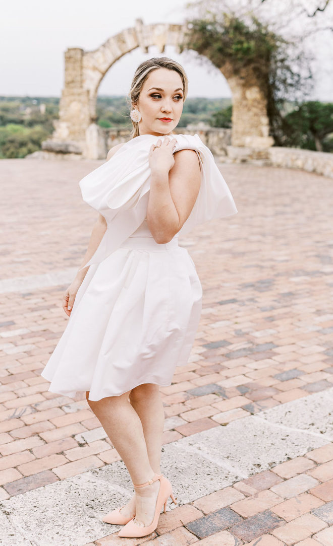 Blonde Bride, wearing white dress with large bow tied on the shoulder and blush high heels, posing on outdoor brick patio at Camp Lucy