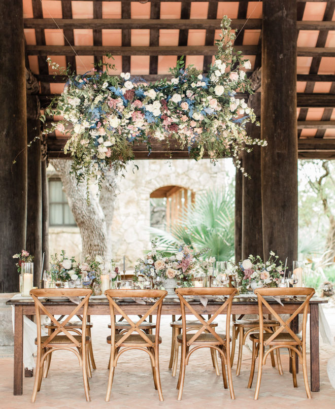 Scarlet Rose Events designed al fresco reception set-up with short farmhouse style table and rustic chic pastel colored florals underneath a hanging wild floral installation at Camp Lucy