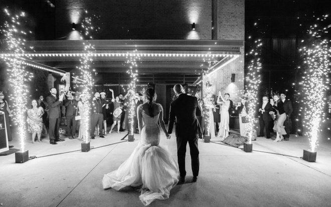 Bride and groom standing hand in hand with large cold sparklers shooting off at either side of them outside the Astorian Houstons, guests are cheering and taking photos using their phones 