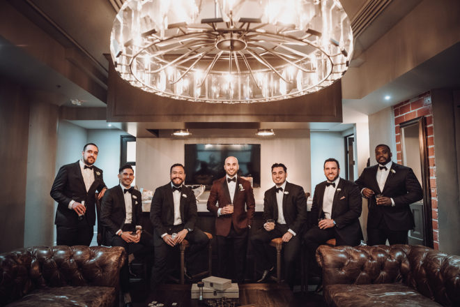 Groomsmen in black tuxedos with black bowties and groom in a maroon jacket with black lapels sit on barstools in the industrial grooms suite fit with chesterfield couches, a statement chandelier and unfinished brick