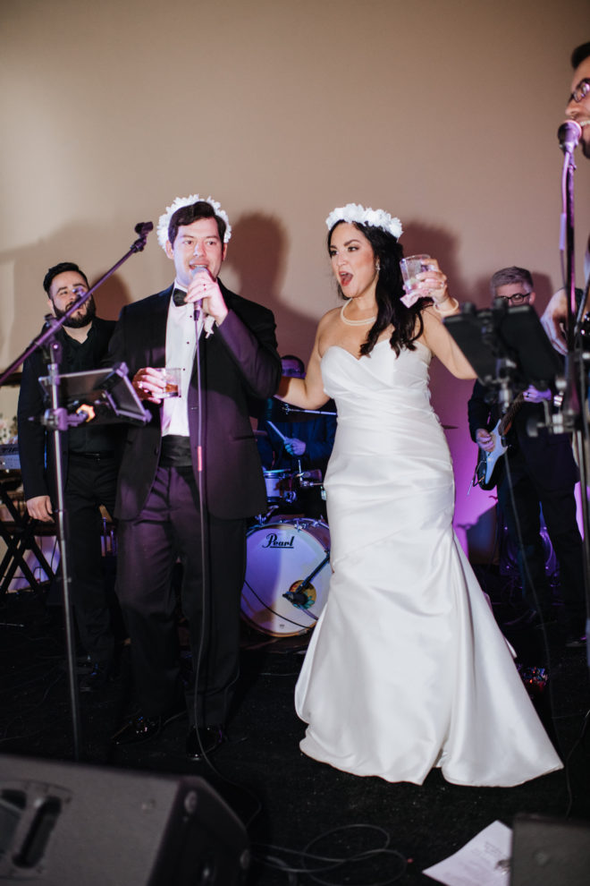 Bride and groom toast in front of band wearing flower crowns. 