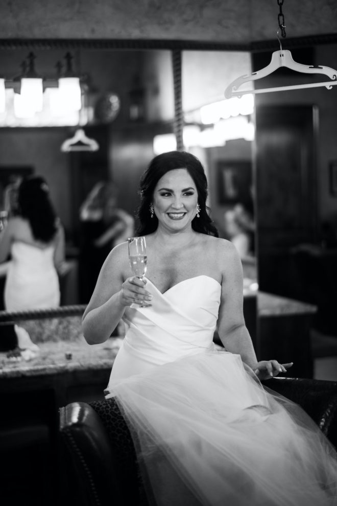 Black and white photo of bride getting dressed in a strapless white gown while drinking champagne. 