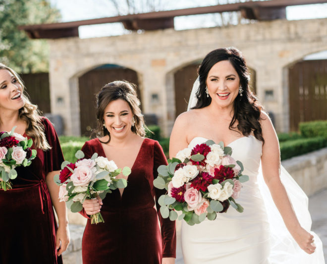 Bridesmaids wearing wine colored velvet dresses and holding roses and greenery bouquets laughing outside with bride who is also holding wine and blush colored rose bouquet outside of Briscoe Manor wedding venue 