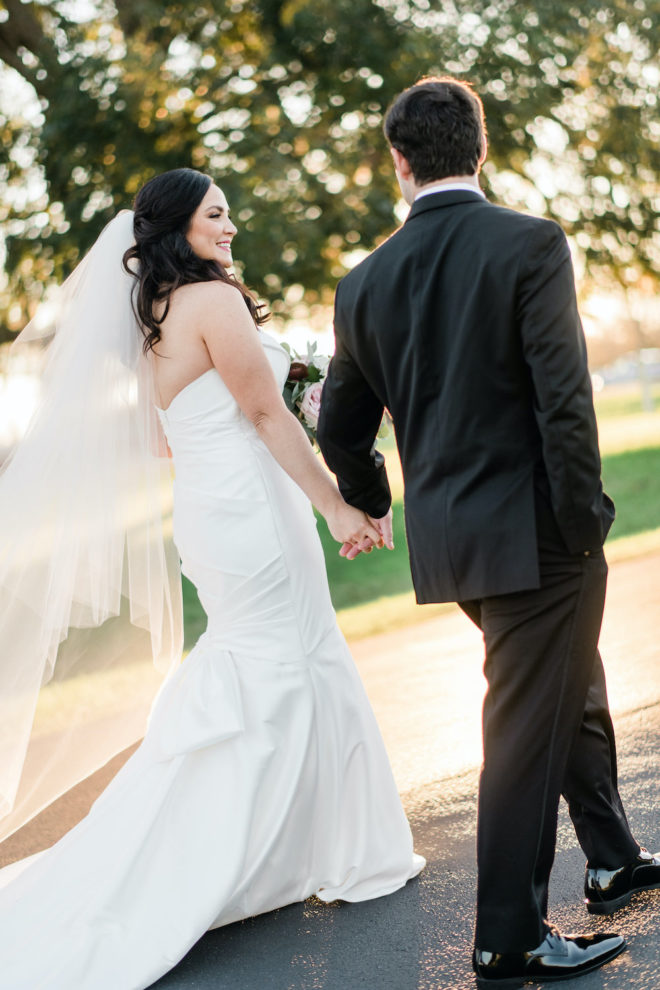 Bride in strapless white dress with veil walking with husband in black tux. 