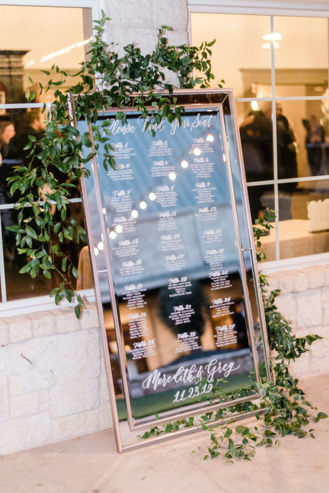 Mirror seating arrangement chart with green florals for reception dinner. 