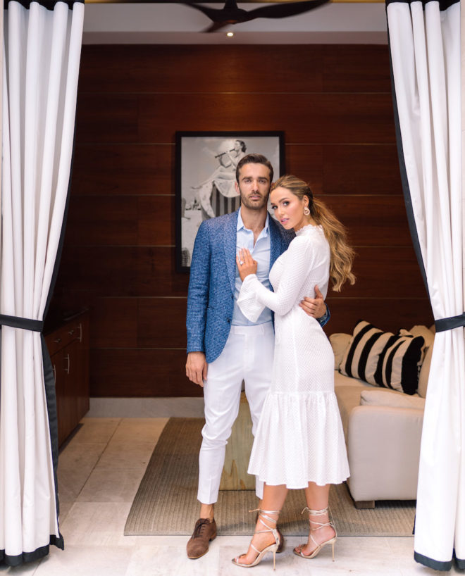 Man in blue suit jacket and white slacks and loafers posing with woman in mid length white dress and stiletto heels inside of luxury pool side cabana at Post Oak Hotel