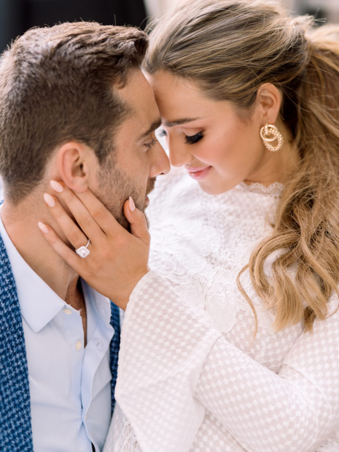 Bride, wearing white textured dress and gold earrings, sitting on grooms lap with her forehead against his 