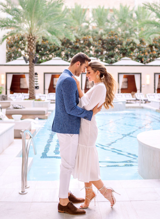 Man in blue blazer and brown loafers holding woman with ponytail, stiletto heels and ankle length dress in front of the Post Oak Hotel's pool and cabanas