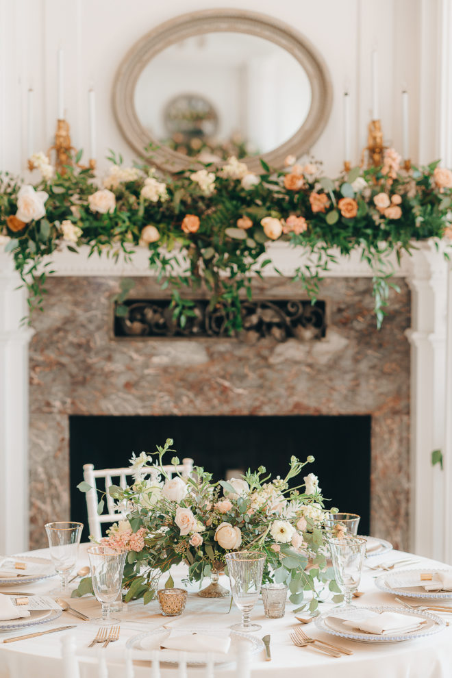 Fireplace adorned with peach and blush florals and greenery behind table set with lush blush botanicals, white linens and blush accents inside of the Mansion at ColoVista