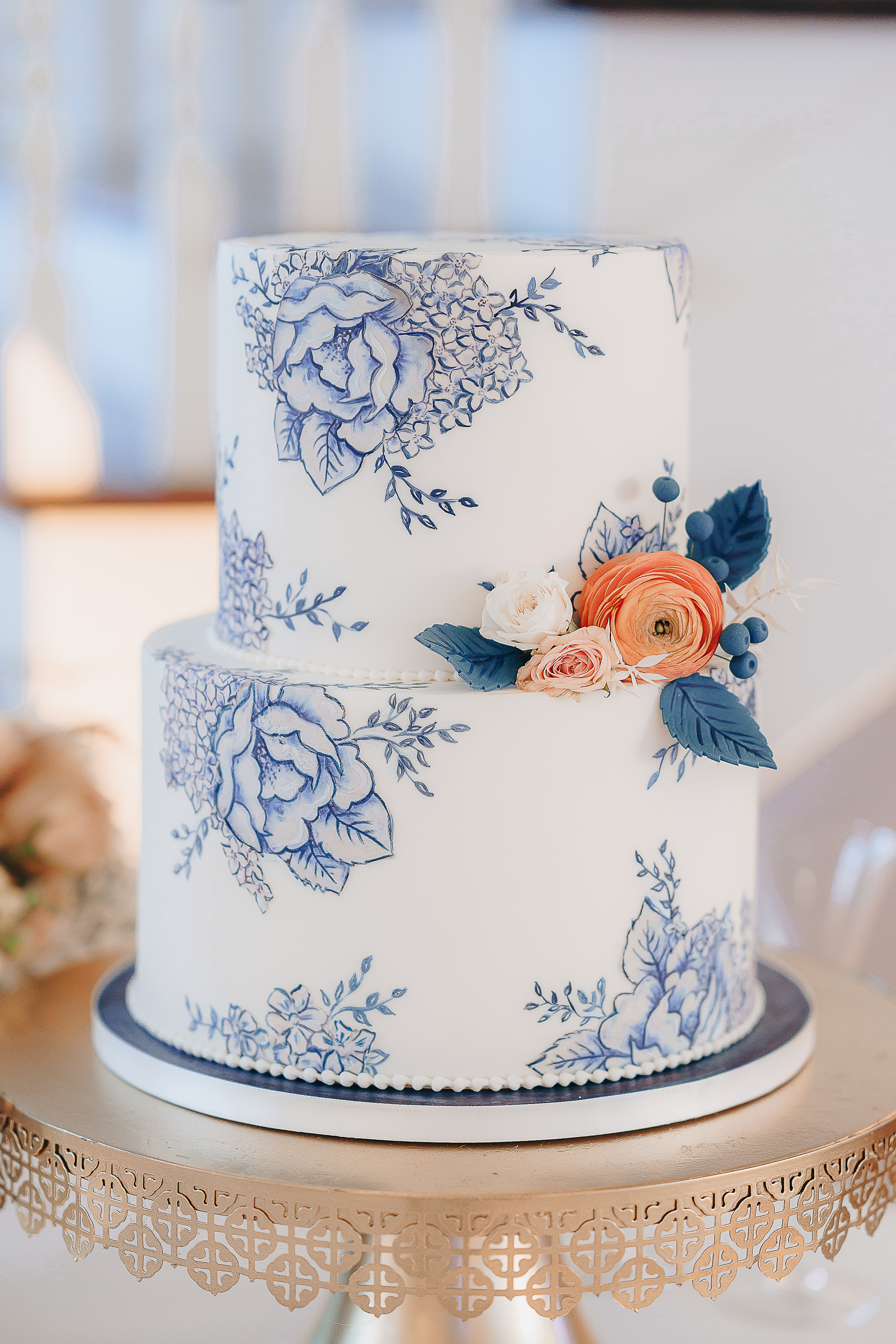 Charming hill country two-tier wedding cake with painted navy flowers atop a gold cake stand.