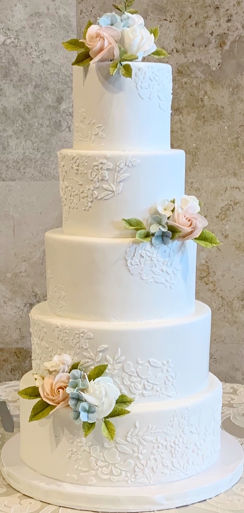 White 5 tier cake with white flower stencil design and light pink and blue 3D flowers.