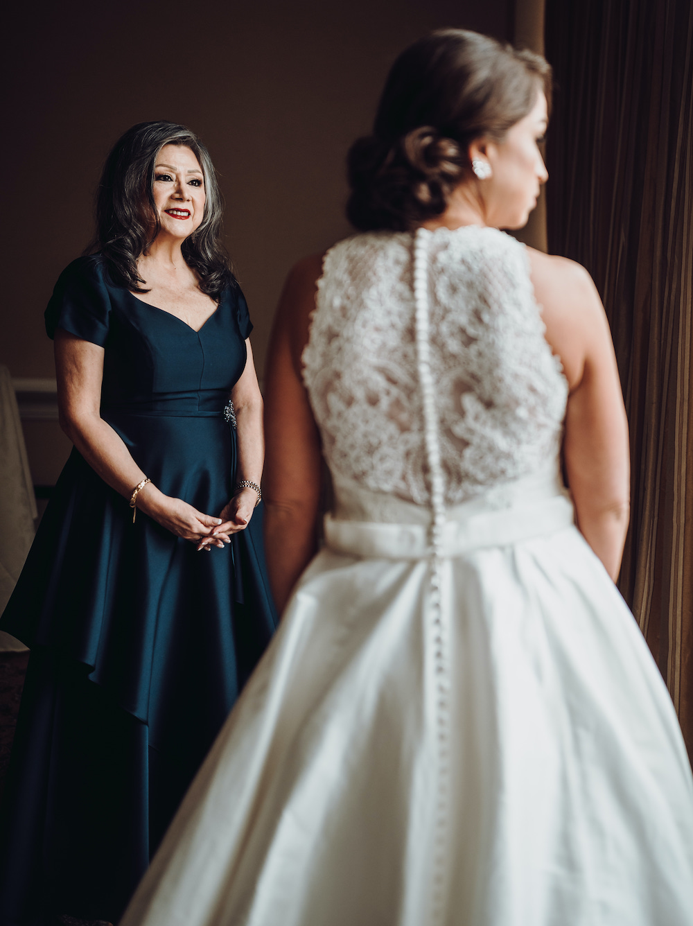 Mother of the bride in black gown smiling at the bride wearing a white wedding gown with lace details on the back of the dress. 