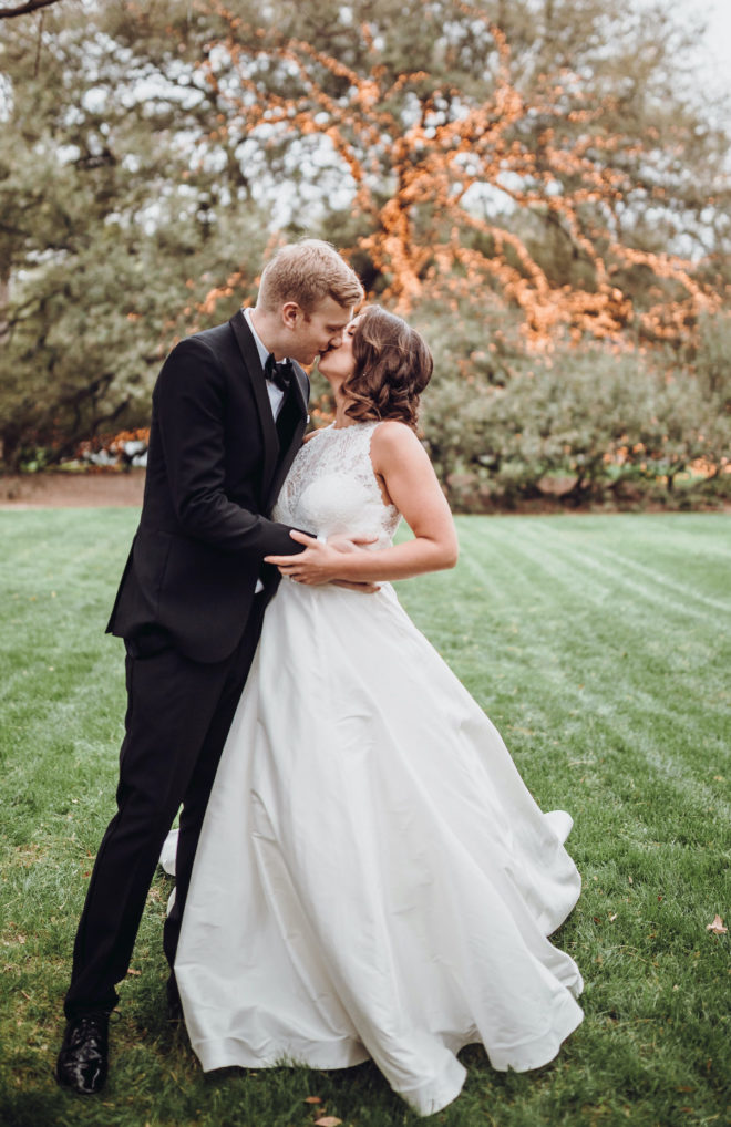 Groom, in black tuxedo and bowtie, kissing bride, in white sleeveless princess cut gown, in front of large Oak tree covered in string lights at the Houstonian Hotel Club and Spa.