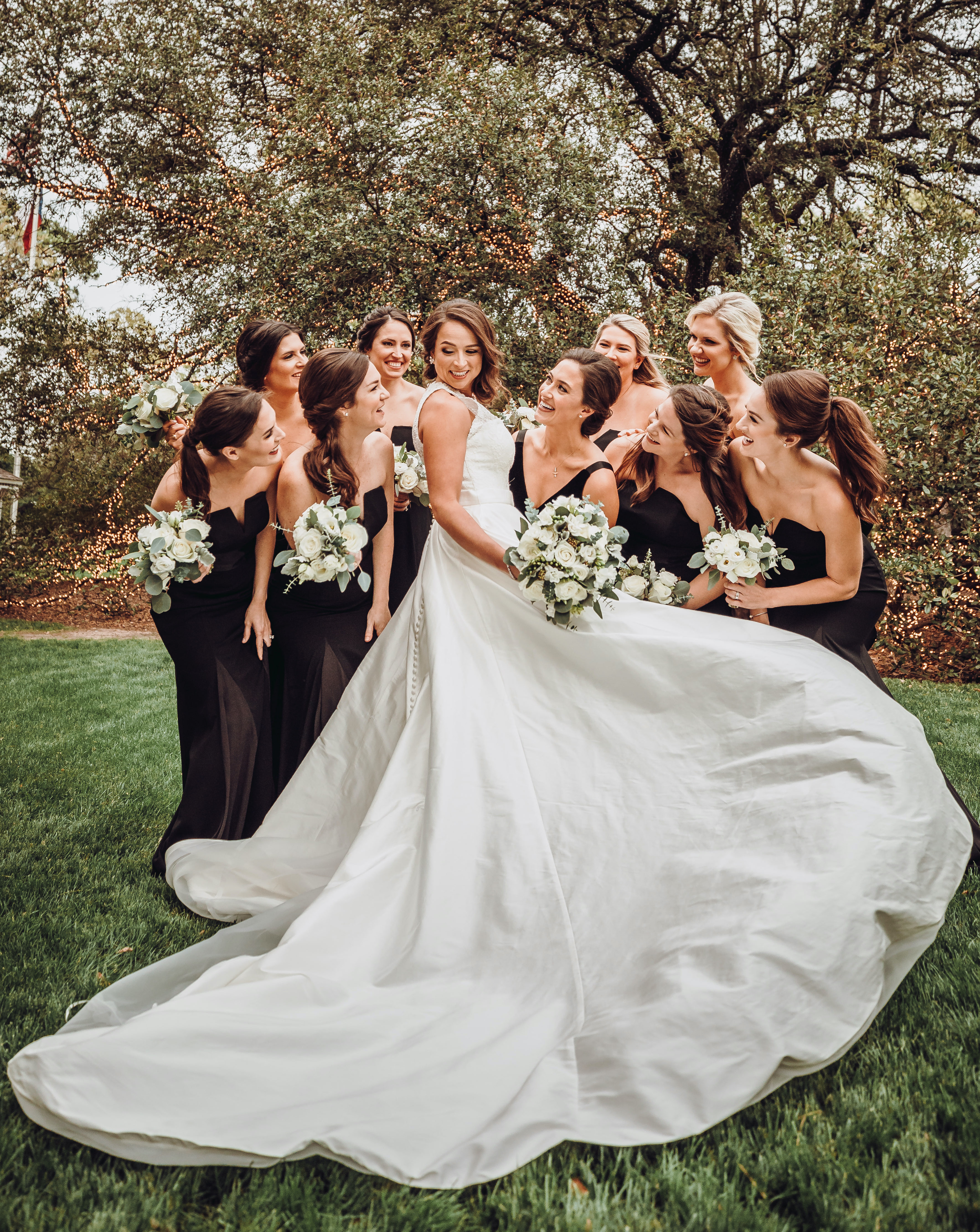 Bride showing off long, elegant wedding gown train as bridesmaids dressed in black admire and smile. 