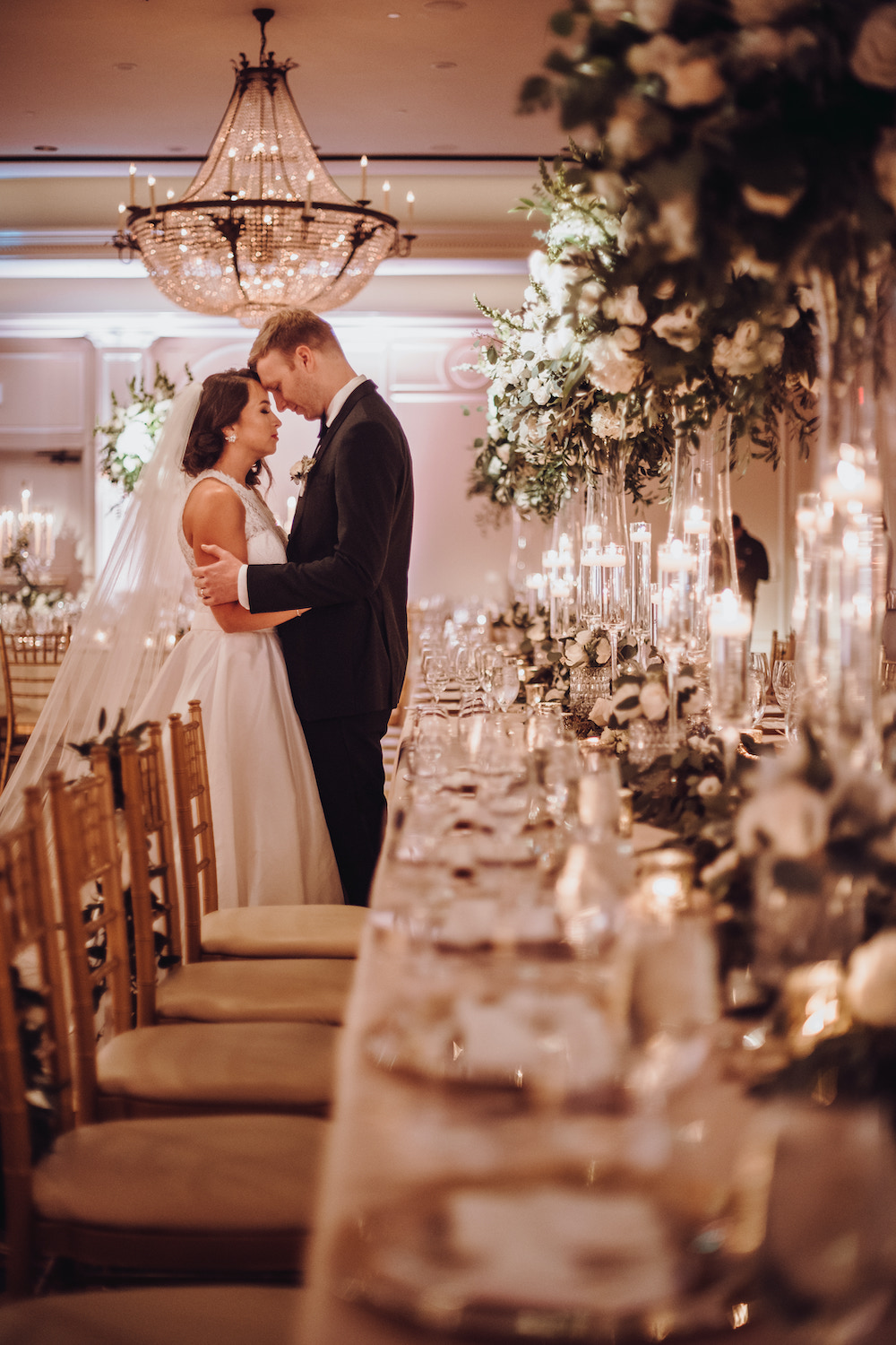 Bride and groom holding each other at the end of long wedding table set with multiple tall blooming floral centerpieces, clear votive holders and strand of lush greenery inside the Houstonian Club and Space ballroom