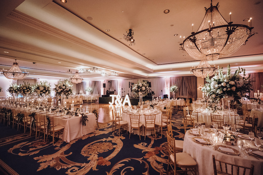 The Houstonian Hotel ballroom decorated for reception with long wedding table adorned with cascading floral centerpieces and round tables with gold chivari chairs and tall blooming white floral centerpieces.