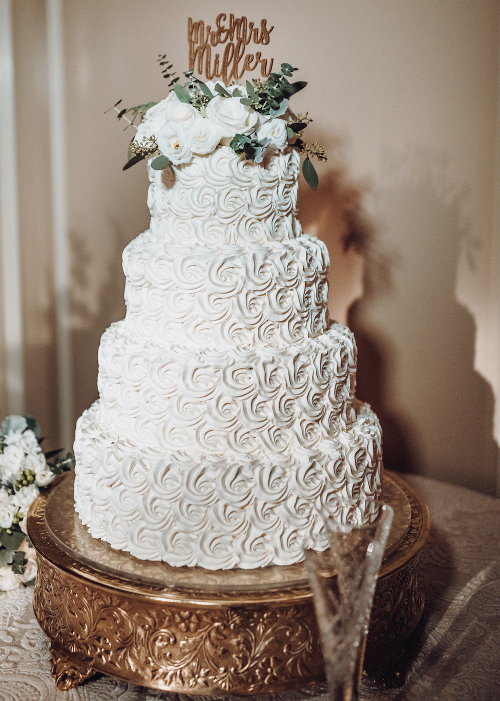 Four tiered white wedding cake by Susie's Cakes with small rose inspired swirls in icing, topped with white roses, eucalyptus and gold lettering which reads, "Mr. and Mrs. Miller" 