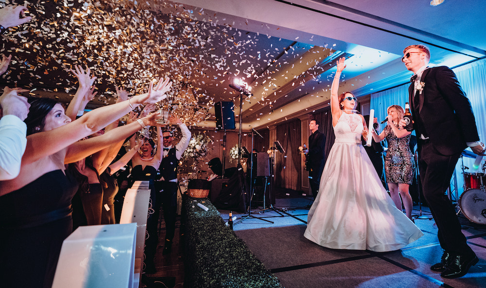 Gold confetti raining down on guests as bride and groom dance on stage with wedding entertainment group. 