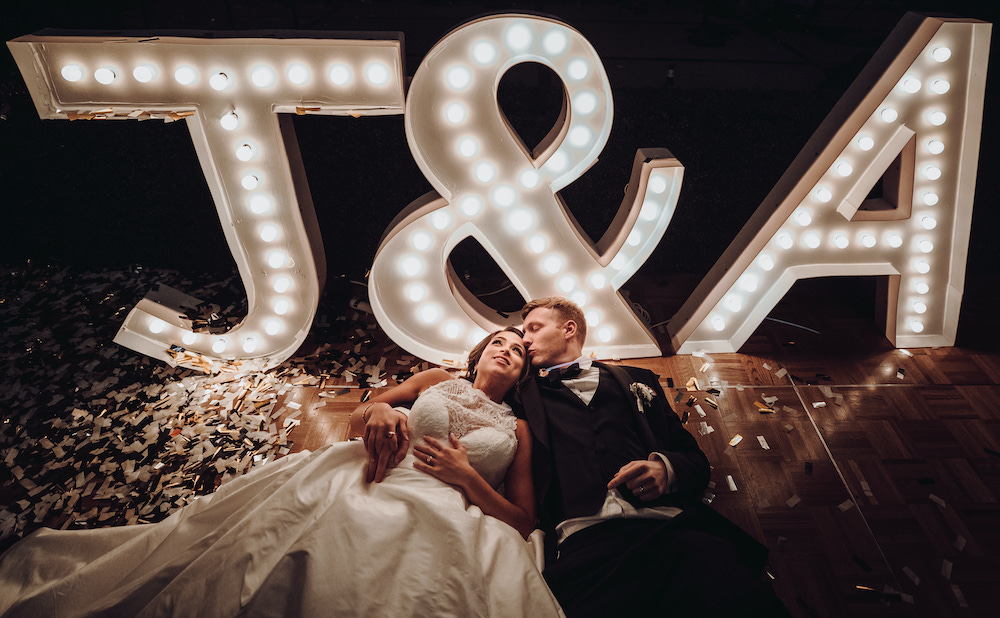 Bride and groom laying side by side surrounded by gold confetti and tall marquee lights reading "J & S"