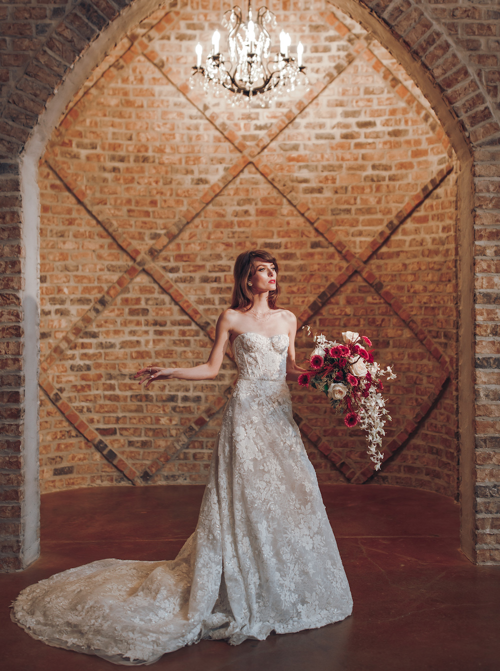 Bride is holding a cascading bridal bouquet with deep tones of red and cream. Her dress designed by Mira Zwillinger is a strapless lace gown that falls to the floor of the wedding venue Iron Manor in Conroe, Texas.