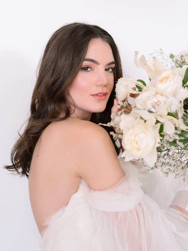 Bride with loosely curled hair, nude makeup and off the shoulder layered white gown holding white monochromatic bouquet of florals 