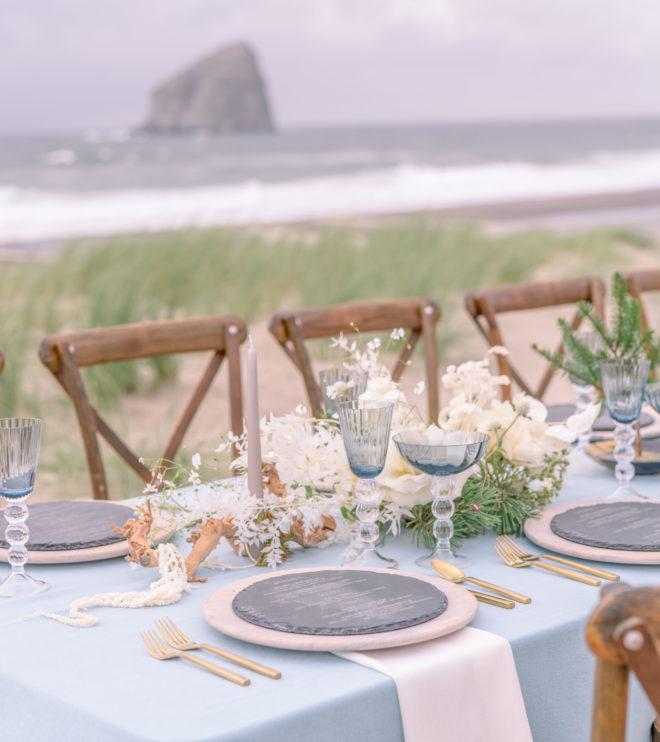 Al fresco tablescape on Cannon Beach- place settings in shades of blue and blush with vintage inspired blue glassware, blush chargers and light grey stone menus.