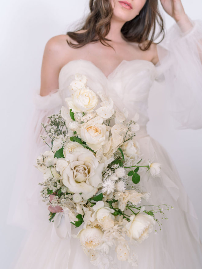 Bride wearing white off the shoulder tulle gown holding cascading bouquet of white and cream monochromatic florals, baby's breath and green botanicals.