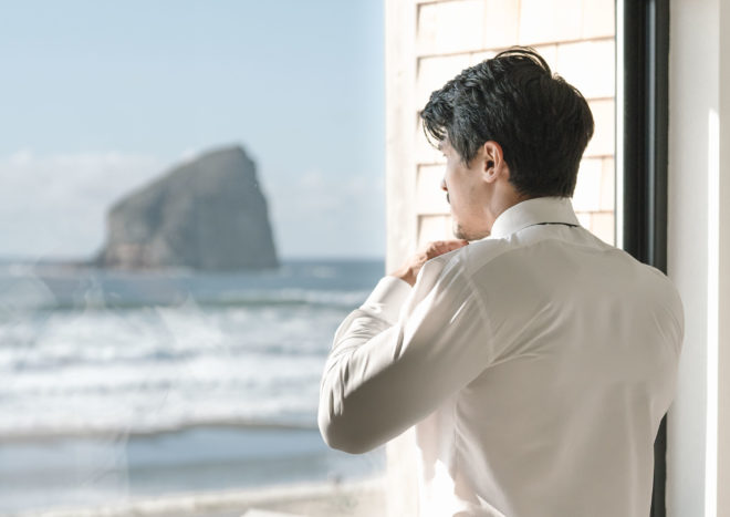 Groom putting his tie before the wedding ceremony, looking out window at Cannon Beach in Oregon