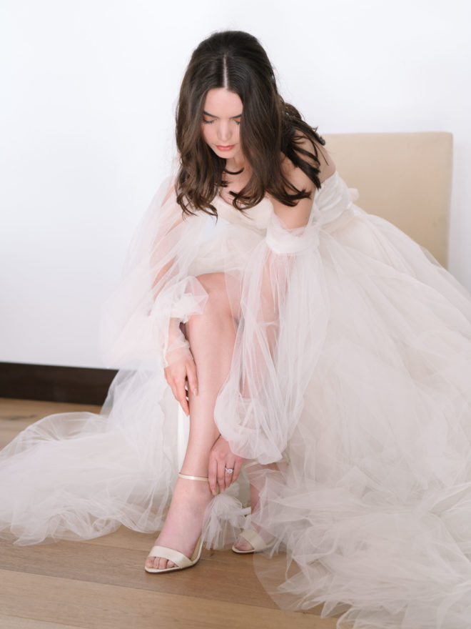 The bride wears a layered tulle wedding dress while putting on white heeled sandals.