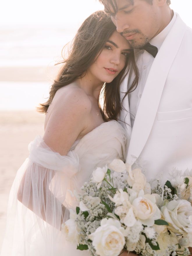 A bride and groom embrace on the beach in Oregon as the sun sets while the bride holds her bouquet.