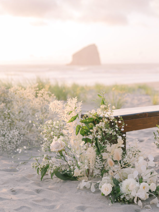 A white wedding floral arrangement sits next to a backless wooden bench at sunset on the beach in Oregon with Haystack Rock in the background.