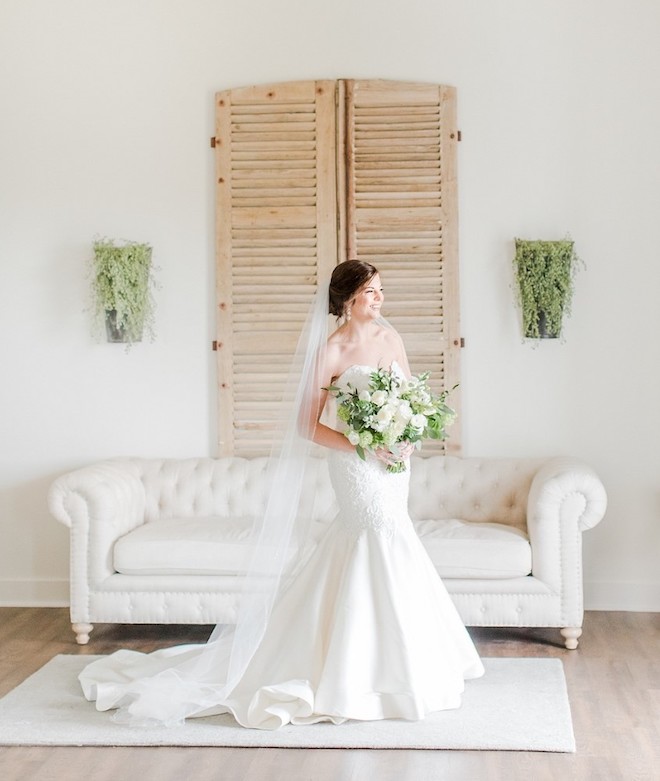 Bride in strappless white gown with train and veil holding green and white bouquet. 