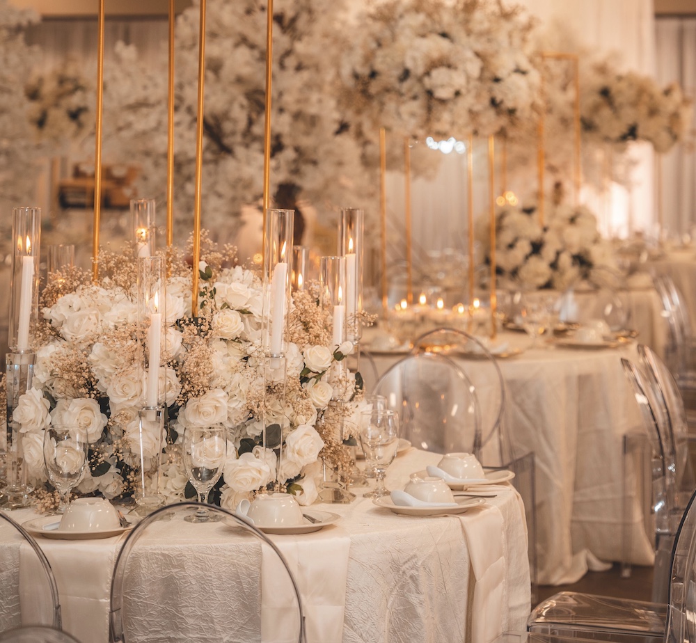 Large white floral arrangement with blush baby's breath and a gold pedestal above, surrounded by acrylic chairs atop white linens by Mod Effect Events.