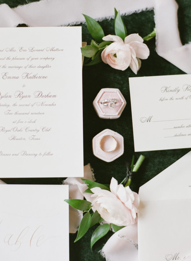 Flay lay of white invitation suite with cursive lettering, two blush rose blossoms and the couple's wedding rings atop pink ring boxes.