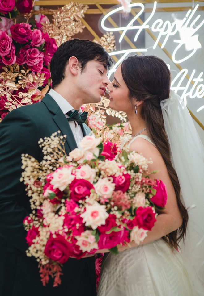 Groom leaning in to kiss the bride, holding a bouquet of blush, fuchsia and ivory silk florals, with LED sign in the background which reads, "Happily Ever After" in ballroom of Hyatt Regency Houston. 