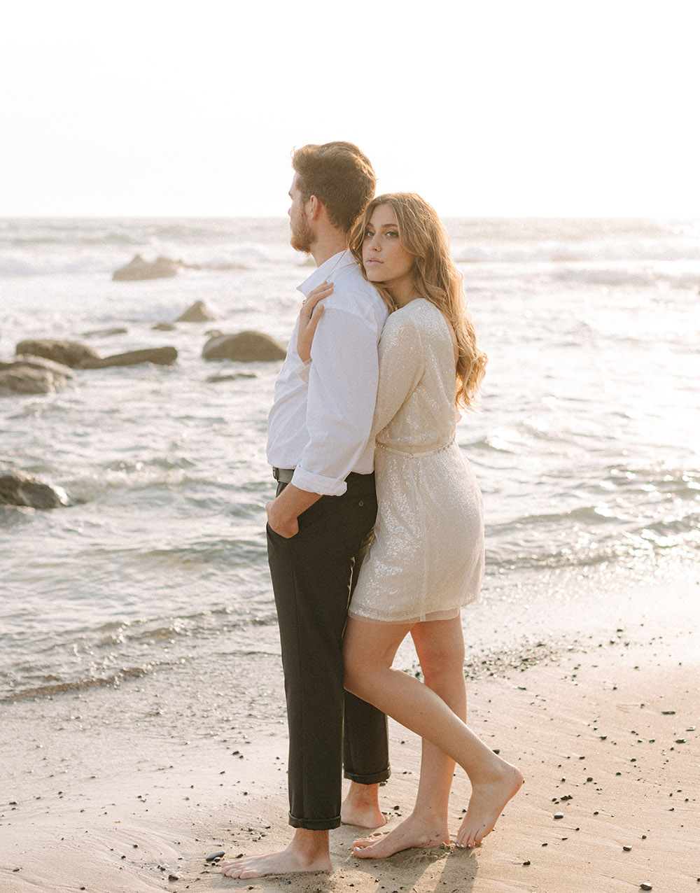 A barefoot bride embraces her groom as the sunlight illuminates the couple and the ocean.