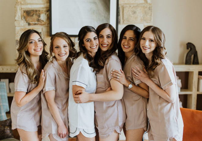 Bride, in white pajamas, poses with five bridesmaids in matching blush pajamas in front of an Austin stone fireplace within a hotel suite at Hyatt Regency Lost Pines Resort and Spa. 