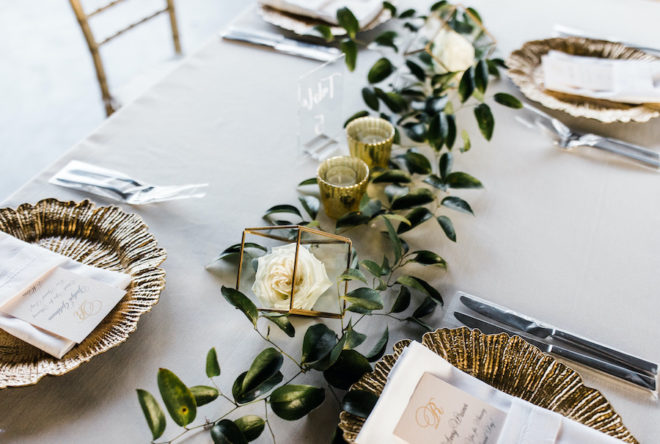 Tablescape with long strand of greenery, gold mercury votives, gold chargers and ivory toned menus tucked into white pressed napkins in the interior event space at Hyatt Regency Lost Pines Resort. 