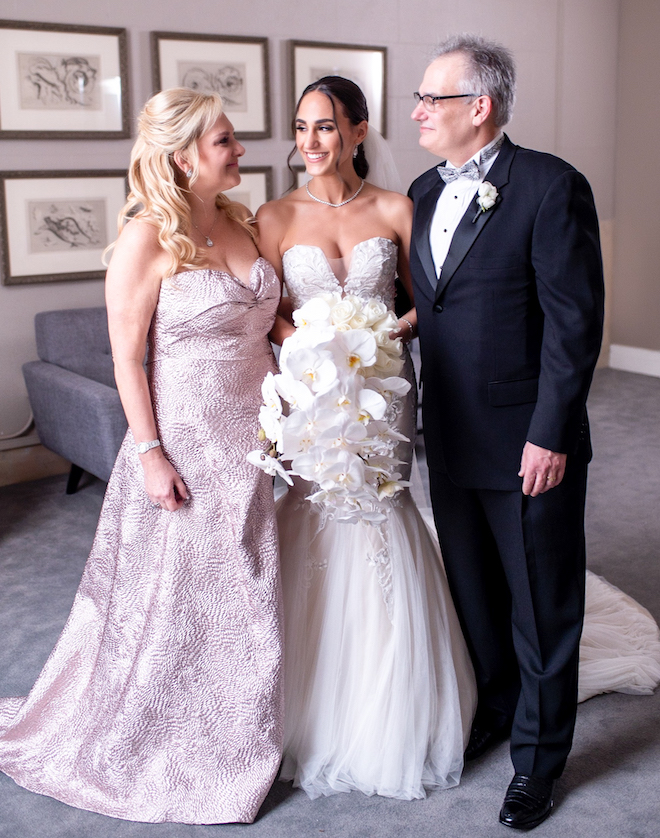 Mother and father of bride look at the bride and smile, as the bride looks to her mother and smiles. 