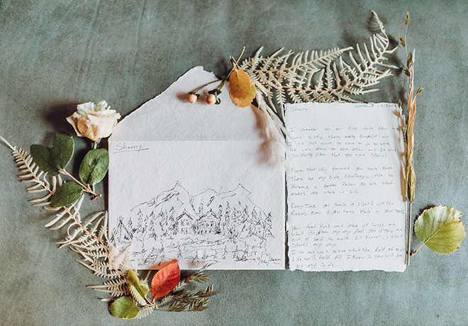 Envelope with hand illustrated picture of woods and mountains with the name "Shanny" on it next to a hand-written letter surrounded by dried florals and leaves.