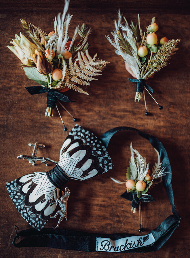 The groom's black and white feather bowtie rests next to dog-shaped cufflinks and wedding boutonnieres of autumn colored botanicals and small grass plumes.