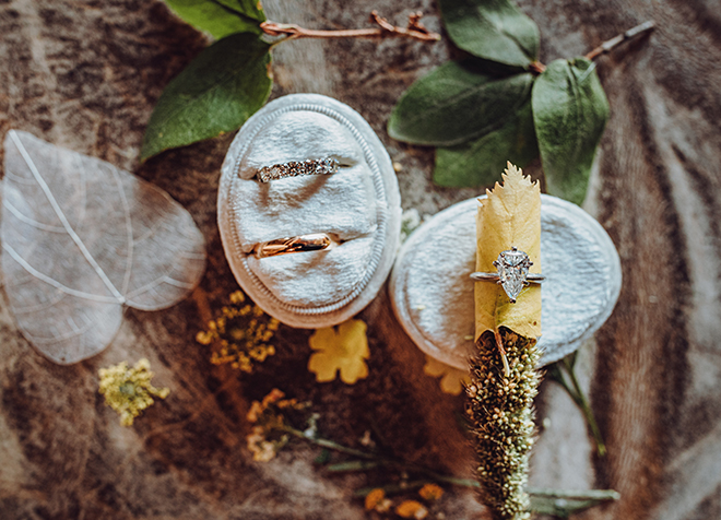 Yellow wildflowers, two green leaves and a white aspen leaf surround two velvet ring boxes holding a diamond wedding band, a solid gold wedding band, and a pear shaped diamond engagement ring. 