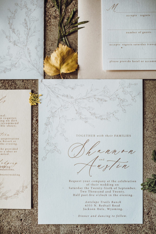 Dried botanicals surround a white and cream colored invitation suite including a white invitation with a light gray floral motif that reads, "Together with their Families Shannon and Austin request your company at the celebration of their wedding"