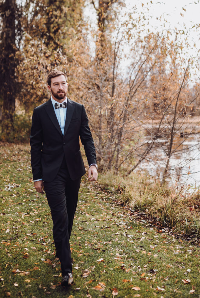 Groom wearing black suit with feather bowtie walks down a leaf strewn grass path aside Snake River at his wedding in Jackson Hole, Wyoming.