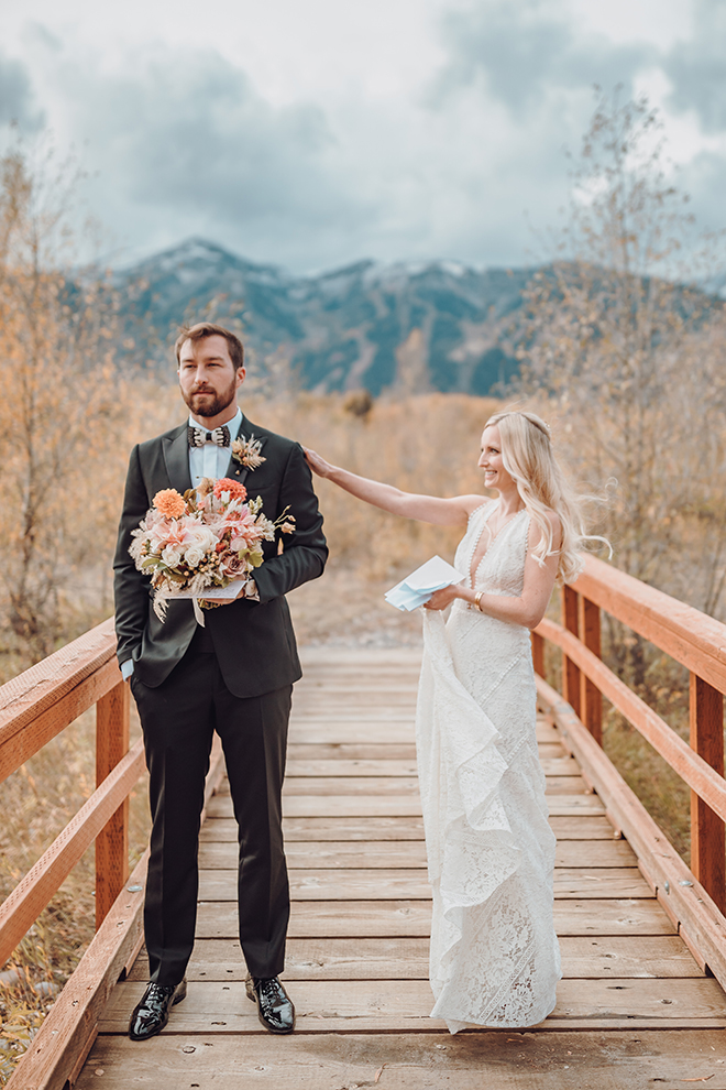 A bride taps her groom on the shoulder before their first look while standing on a bridged with the Teton mountians in the background.