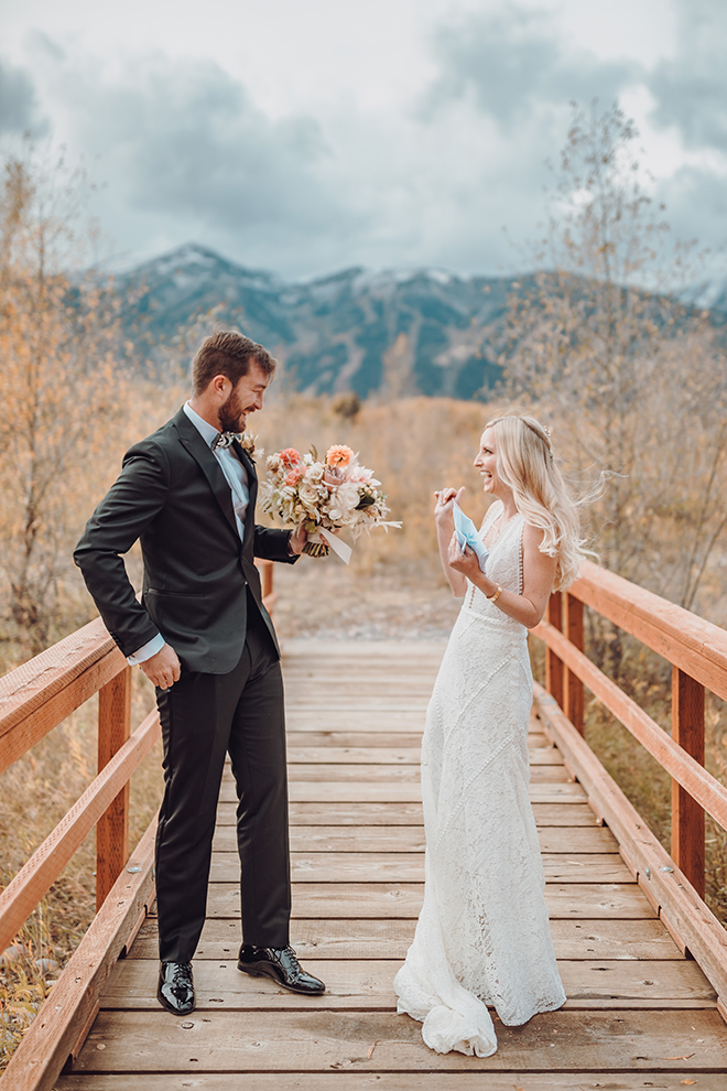 Groom in black suit with blue undershirt holding bouquet of florals smiles at a smiling bride in a long lace fitted gown while standing on an outdoor bridge with the Grand Teton mountains behind them in Jackson Hole, Wyoming. 