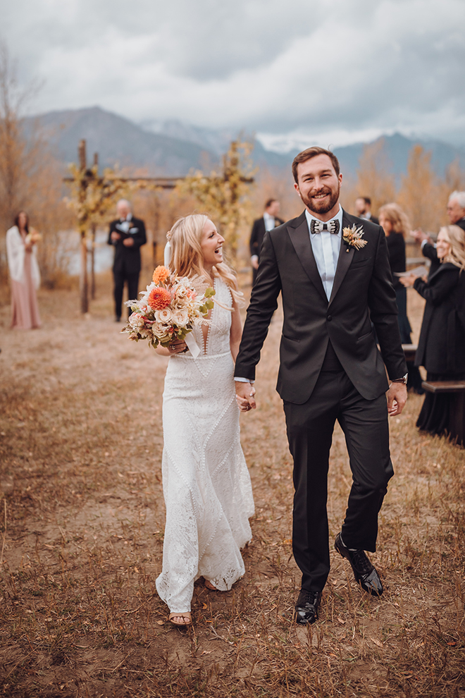 A bride and groom walk hand in hand after their wedding ceremony with an aspen-adorned altar and the Teton mountains in the background as their guests clap.